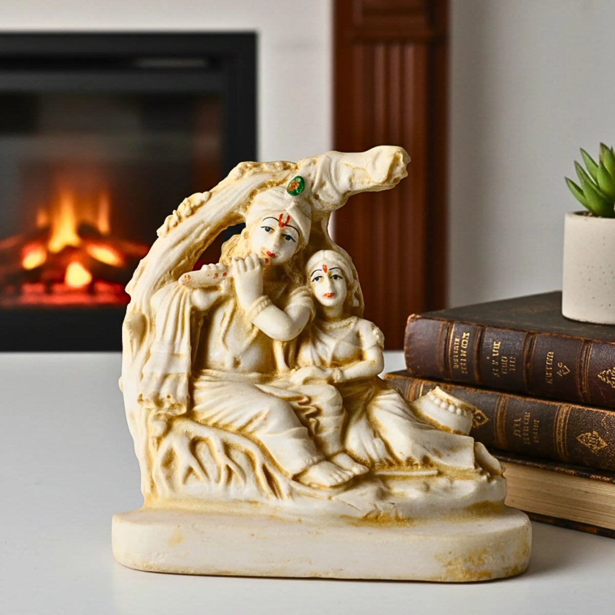 An off-white stone or resin figurine of a Radha Krishna under a tree, displayed on a tabletop with a blurred fireplace and books in the background.