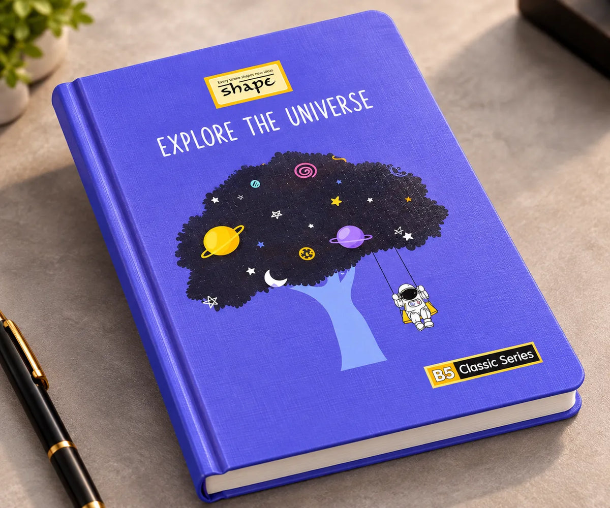 A blue hardcover notebook titled 'EXPLORE THE UNIVERSE' from the 'BS Classic Series' by 'shape', featuring a space-themed illustration of a tree with planets and an astronaut. The notebook is photographed on a gray textured desk next to a black pen.