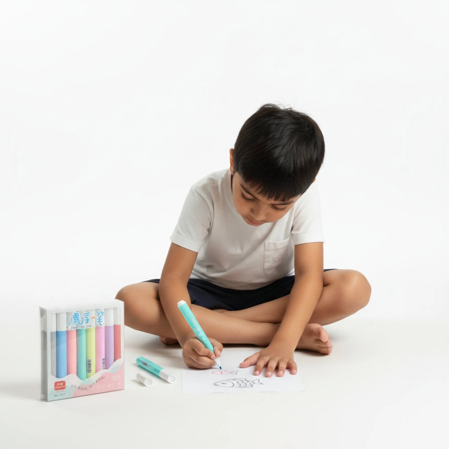 Child drawing with colorful markers on a white surface, with a marker set in the foreground.