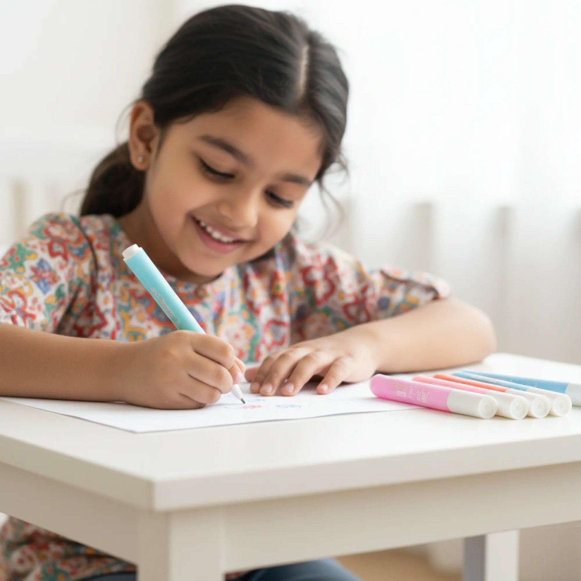 Young girl writing with a pen at a table, surrounded by colorful markers.