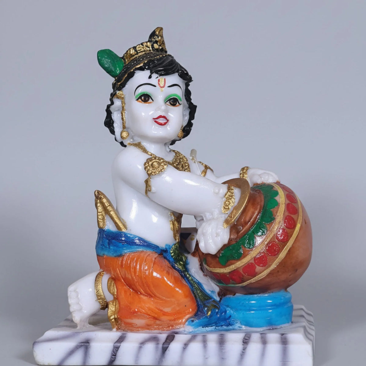 A hand-painted ceramic figurine of a child deity, seated on a white marble-like base and holding a decorated pot, shown against a plain light gray background.