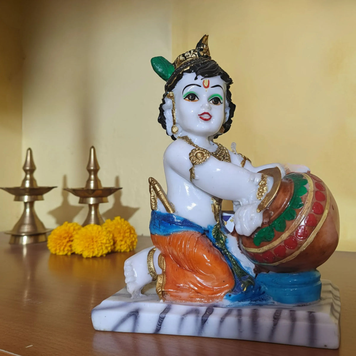 A colorful figurine of a child deity seated and holding a decorated pot, placed on a marble-like base on a wooden surface with brass oil lamps and yellow flowers in the background.