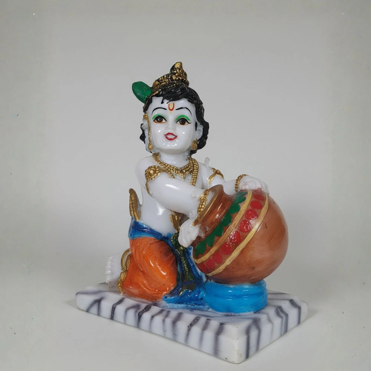 A colorful hand-painted figurine of a child deity playing a drum, seated on a marble-like base against a plain light gray background.