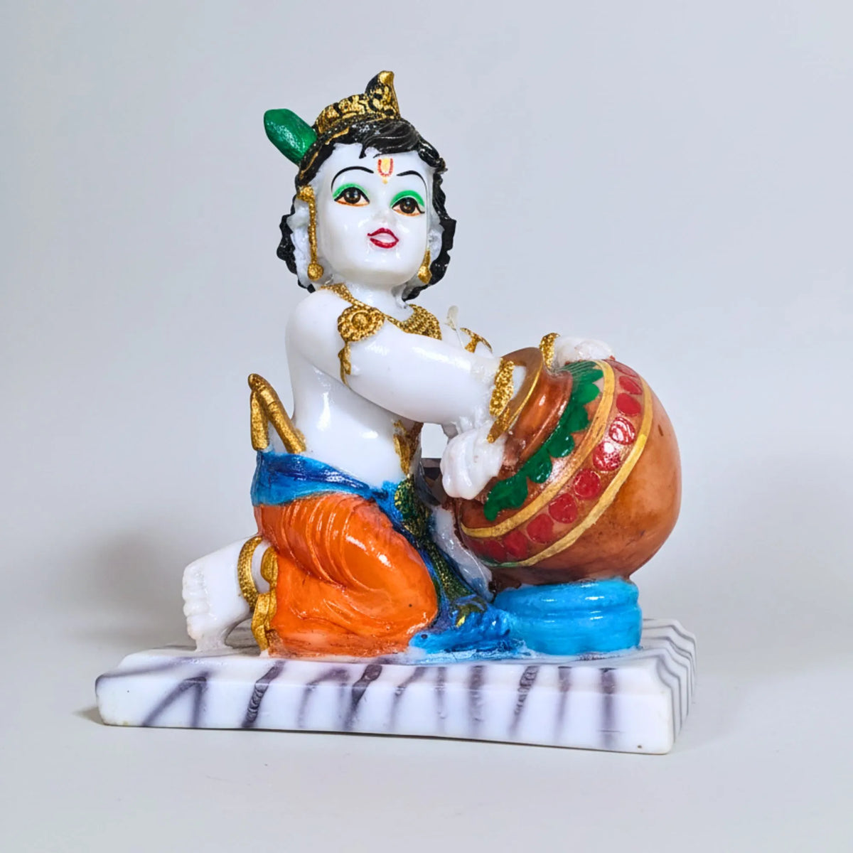 A colorful figurine of a seated child deity holding a decorated pot, shown on a plain light gray studio background.