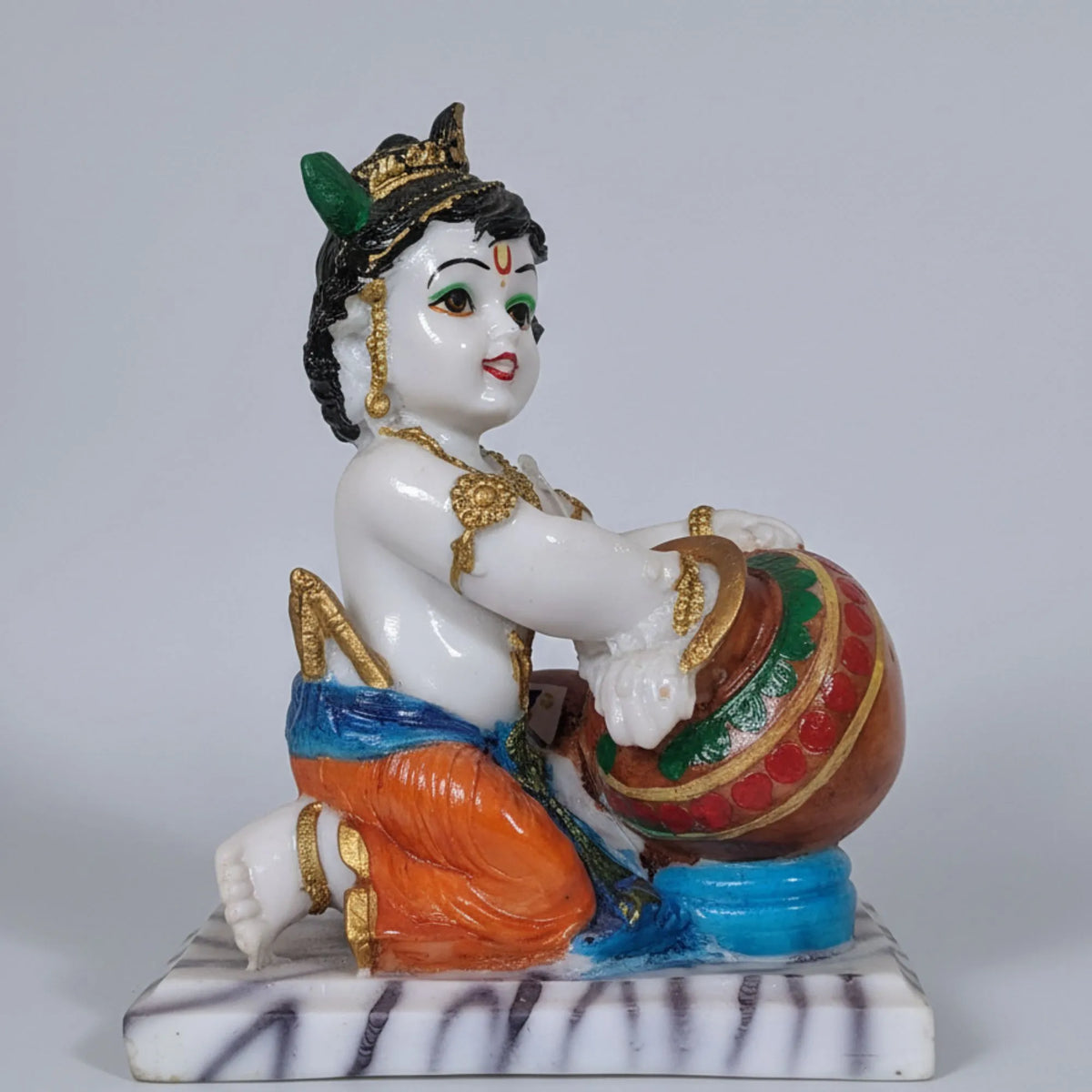 A hand-painted ceramic figurine of a child playing a drum, set against a plain light gray background. The figurine features a white body, orange and blue lower garment, gold jewelry, and a green and red patterned drum, mounted on a white marble base with black zebra-like stripes.