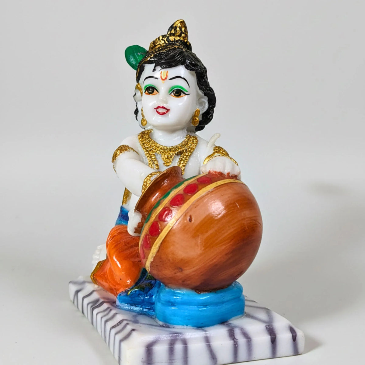 A hand-painted resin figurine of a child playing a drum, set against a plain light gray background. The figurine features a white face and hands, black hair, a gold crown and jewelry, orange clothing, and a blue base.