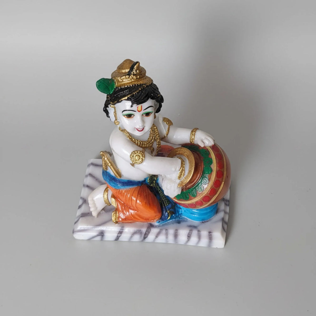 A small decorative figurine of a seated child playing a drum, shown on a plain light gray studio background. The figure has a white face and upper body, dark hair, gold jewelry and crown details, an orange garment, blue lower garment accents, and a marbled white-and-gray base.