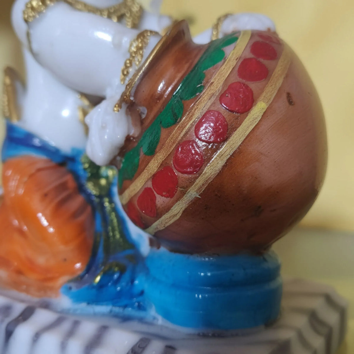 A close-up of a colorful figurine showing a white hand holding a brown pot decorated with red circular motifs, green leaves, and gold trim, set against a soft yellow background.