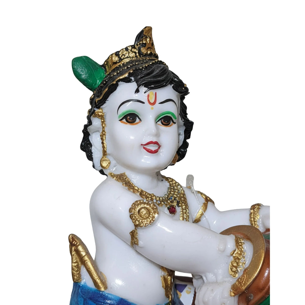 A close-up of a decorative figurine of a child deity with a white face and body, black hair, a gold crown and jewelry, green and red facial details, and blue fabric at the bottom, shown against a plain white background.