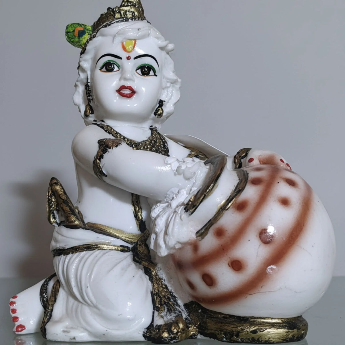 A white porcelain figurine of a child deity with gold jewelry and a red bindi, kneeling beside a large decorated pot on a plain light gray background.