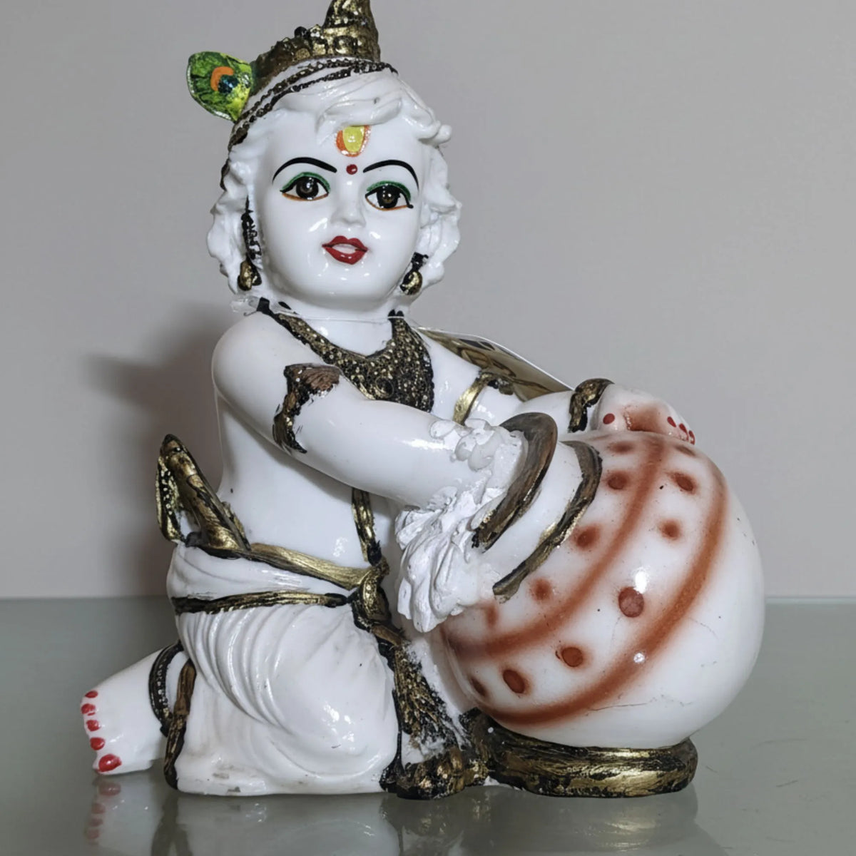 A white ceramic figurine of a child deity holding a decorated pot, set against a plain light gray background.