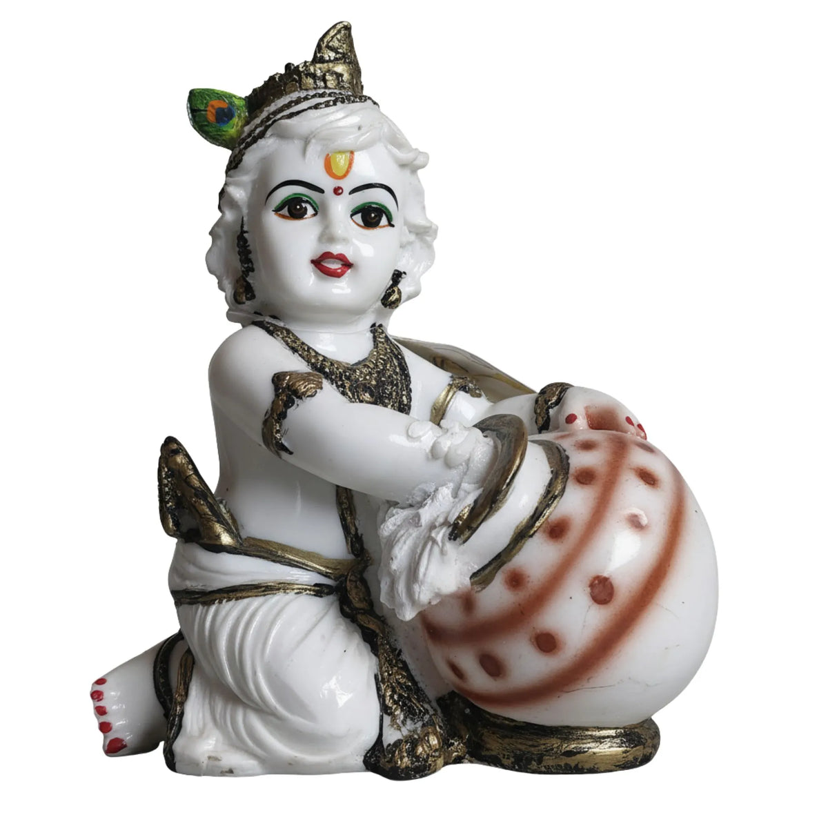 A white porcelain figurine of a child deity, likely Krishna, kneeling and playing a decorated drum. The figurine features gold accents, a peacock feather in its hair, and red painted details on the face and drum.