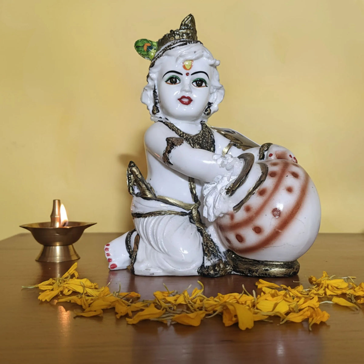 A white Lord Krishna figurine holding a drum, styled on a wooden surface with a burning brass diya and yellow flower petals against a yellow background.